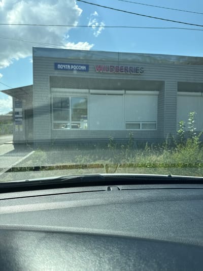 An exterior view of a Wildberries and Pochta Rossii (Russian Post) storefront is captured through a vehicle's windshield, which is dirty and distorts the image. The building is modern with a gray, corrugated metal facade. The Wildberries logo, in pink and purple gradient lettering, is prominently displayed above the entrance. To the left of the Wildberries logo, in a blue rectangular sign, is the text "ПОЧТА РОССИИ". The storefront has large windows, some covered with white roller shutters. Green vegetation is visible in front of the building. The scene appears to be in Romanovskaya, Russia, based on the presence of the Pochta Rossii sign, a state-owned postal service. The foreground shows the interior of a car, including the dashboard and windshield wipers. The sky is partly cloudy with blue sky visible.
