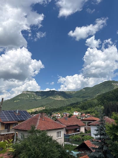 The image depicts a village nestled in a valley with rolling green mountains in the background under a partly cloudy blue sky. In the foreground, several houses with red tile roofs are visible. One house prominently features solar panels on its roof. Green trees and foliage surround the houses, creating a lush, natural environment. The scene appears to be a residential area within the village of Zgorigrad, Bulgaria, known for its natural beauty and mountainous terrain.