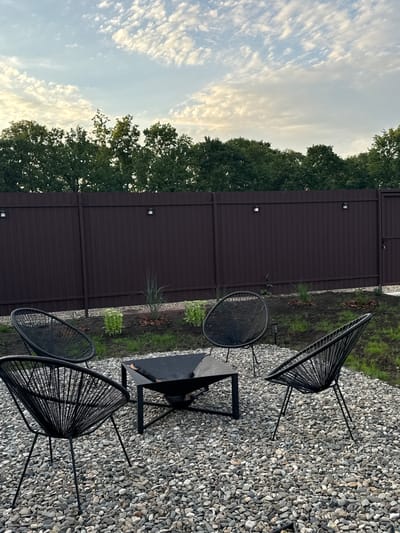 In the city of Starokorsunskaya, Russia, a modern outdoor seating area is depicted. The scene is set on a gravel patio, surrounded by a dark brown metal fence. The sky above features scattered white clouds against a pale blue backdrop. Four black wire-frame chairs, in a distinctive Acapulco style, are arranged around a black metal fire pit. The fire pit has a square top and a geometric base. Beyond the seating area, the ground transitions to soil and grass, with small plantings of greenery and mulch. The fence is punctuated by several small, square outdoor lights. The overall impression is of a contemporary, minimalist outdoor living space.