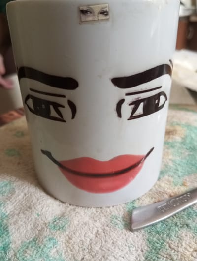 The image shows a white mug decorated with a face. The face has large, dark eyebrows, cartoonish eyes, and red lips. Above the eyes, there is a small rectangular sticker with an image of eyes. The mug is sitting on a textured, light-colored cloth with faint green and blue patterns. In the lower right corner, a sliver of a metallic object, possibly a utensil, is visible. The background is blurred, but suggests an indoor setting. The location in the city of Dymer, Ukraine is not determinable from the image.