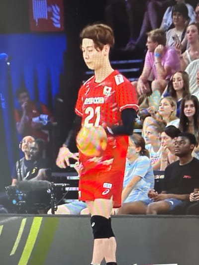 In the media file, a crowd of people watches a volleyball match. The primary subject is a volleyball player in a red uniform with the number 21 on their back, who is in mid-action, potentially serving or receiving. The player is wearing black shorts and arm sleeves. The background shows other players and a seated audience. The scene appears to be indoors, likely a gymnasium, indicated by the lighting and the presence of bleachers. The mood seems to be one of focused attention and possibly excitement from the spectators. Some audience members are looking towards the court, while others are captured in candid moments. The lighting suggests it could be daytime or evening, but there are no clear indicators of weather. Location cues for Peremyshliany, Ukraine, are not visible within the frame. Visible text on the player's uniform includes "RCP COM" and the number "21." There are also some unidentifiable logos on the uniform. The overall image captures a moment of athletic competition and spectator engagement.