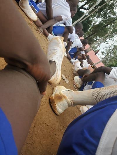 The image depicts a group of young individuals, likely students, gathered outdoors on a dirt surface. They are seated, with some leaning against each other. The individuals are wearing matching white t-shirts and blue shorts, suggesting a school uniform or team attire. Several are wearing white athletic shoes, some of which appear dirty. The environment includes trees and a red-colored building structure in the background, indicating an outdoor setting, possibly a schoolyard or sports facility in Bahit, Nigeria. The scene conveys a sense of rest or waiting.