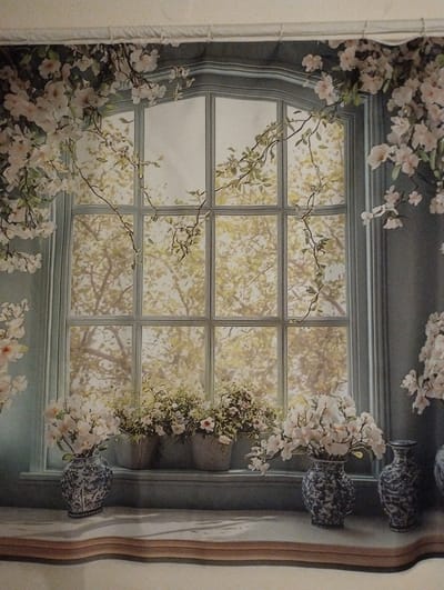 This is a detailed, flat image, possibly a fabric print or a painted mural, depicting a romanticized scene of a window with floral arrangements. The focal point is a large, multi-paned window with an arched top, framed by a light blue-grey structure. Outside the window, a soft, out-of-focus image of trees with light yellow-green foliage suggests a sunny day.
Draped over the window frame and surrounding wall are abundant branches of delicate white and pink blossoms, with some green leaves. These flowers cascade down from the top and sides, creating a lush, organic frame.
On the windowsill below the window, there are several floral arrangements in decorative vases and planters. On the left, a blue and white patterned vase holds a bouquet of white flowers. To its right, there are two grey planters filled with more white flowers and greenery. Further to the right, another blue and white patterned vase holds a different arrangement of white flowers. The windowsill itself is a light-colored surface with a subtle, wavy texture. The background wall on the sides of the window is a muted blue-grey.
The overall impression is one of a peaceful, perhaps slightly nostalgic, domestic setting, emphasizing natural beauty and floral motifs. There is no indication of any specific location within the city of Municipal Unit of Patras, Greece, or any people or actions taking place.