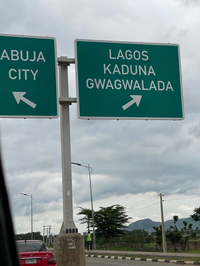 An eye-level, medium outdoor shot features two green directional highway signs against a cloudy sky. The sign on the left reads "ABUJA CITY" in white capital letters and has a white arrow pointing left. The sign on the right reads "LAGOS KADUNA GWAGWALADA" in white capital letters and has a white arrow pointing right. Both signs are mounted on a gray pole. In the background, a red car is parked on the right side of the road. A person in a yellow safety vest stands on the sidewalk. The surrounding environment includes trees, mountains in the distance, and power lines. The scene appears to be located near Immigration Quarters, Nigeria.