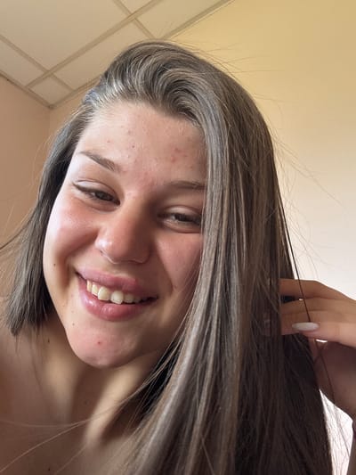 This media file is a close-up, low-angle selfie of a young woman. Her head is tilted to the side, and her hair is long and brown with some lighter streaks, partially obscuring her face and framing her head. A hand with long, white fingernails is visible at the top of the frame, gently touching her hair. The woman is smiling, revealing her teeth, and her eyes are open and looking towards the camera. Her skin appears slightly oily, with some small blemishes and redness on her cheeks and forehead. The background is a plain, light beige wall with some horizontal and vertical lines, suggesting an indoor setting, possibly a room with tiled walls or ceiling panels. The lighting is soft and appears to be natural, coming from above and slightly to the side. There is no visible text in the image. The overall mood is casual and happy. The location context is Prodanovtsi, Bulgaria, though this is not directly evident from the visual cues in the image itself.