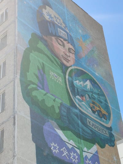 A mural painted on the side of a multi-story residential building in Kirovsk, Russia, depicts a young person wearing a hat and gloves, holding a circular emblem. The mural is vibrant, with the central figure dressed in a green jacket and a blue and purple patterned scarf or garment. The hat features a snowflake design. The circular emblem held by the person contains a miniature winter landscape, including snow-covered mountains, a yellow vehicle, and a building. The word "КИРОВСК" (Kirovsk) is prominently displayed on a banner beneath the emblem, indicating the city's location. The background of the mural includes a stylized star and a spectrum of colors, suggesting a sky or aurora. The building itself is made of concrete and has visible windows, with the mural covering a significant portion of its exterior wall. The sky in the upper right corner is a clear blue.