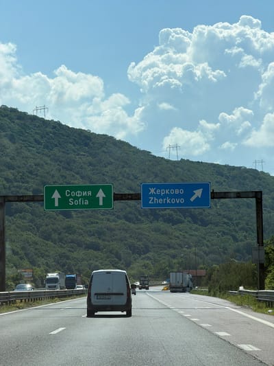 The image shows a highway in Potop, Bulgaria, with a mountainous, forested background under a cloudy sky. Two overhead road signs are visible. The green sign on the left indicates "Sofia" with upward arrows, meaning straight ahead. The blue sign on the right indicates "Zherkovo" with a right-pointing arrow, suggesting a turn or exit to the right. Various vehicles, including trucks, vans, and cars, are traveling on the multi-lane highway. The terrain is characterized by steep, tree-covered slopes, with power lines visible on the hilltops. The lighting suggests it is daytime.