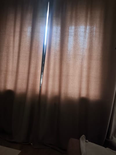 The image shows a close-up view of a tan-colored curtain, with a vertical slit in the fabric. Bright light streams through the opening, casting vertical shadows on the curtain. The bottom of the image shows a dark floor and the edge of a light-colored piece of furniture, possibly a sofa or chair, with a white fabric draped over it. There is no visible indication of the city of Syktyvkar, Russia.