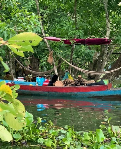 In the city of Soutr Nikom, Cambodia, a long, narrow boat, painted red and blue, is positioned on calm, green water. The boat contains several people and various objects. In the front, a person wearing a dark hoodie is seated, facing away from the camera, and appears to be holding a brown bag. To the left of this person, two blue containers and a white container are visible. Further to the left, a person in a light-colored shirt is seated, also facing away from the camera, near the edge of the boat. In the rear of the boat, there are several logs and other items, including what appears to be a yellow and green bag. The boat is surrounded by lush green foliage and trees, with hammocks and draped fabric visible in the background, suggesting a relaxed, outdoor environment. The water is partially covered with green aquatic plants.