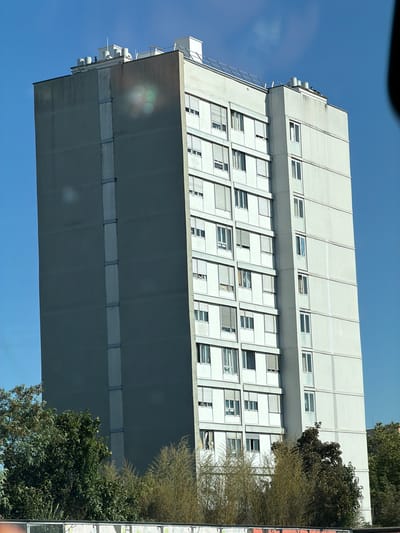 An exterior, medium-range, eye-level shot captures a tall, grey residential apartment building in Bron, France. The building features a light-colored facade on its right side, punctuated by numerous windows, many equipped with white shutters. The left side of the building is a uniform grey with a vertical band of metal fixtures running its height. The top of the building is flat and includes several ventilation units and what appears to be a solar panel array. The sky is a clear, bright blue, with some lens flare visible on the grey section of the building. Trees and foliage are present at the base of the building, obscuring the lower levels. The overall impression is of a functional, urban residential structure under a sunny sky.