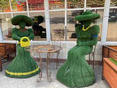 This image depicts an outdoor seating area, likely a cafe or restaurant, in the village of Beskol Rural District, Kazakhstan. The focal point is two topiary figures, sculpted to resemble women dressed in large hats and floor-length gowns. The figures are covered in green artificial grass, with yellow and blue accents on their attire. One figure on the left is holding a yellow purse. A wrought-iron table with intricate scrollwork stands between the two figures. In the background, a building with large windows is visible, reflecting the street outside. Wooden benches and planters are also part of the scene. The ground is paved with interlocking concrete tiles. The overall impression is an artistic and whimsical outdoor decoration.