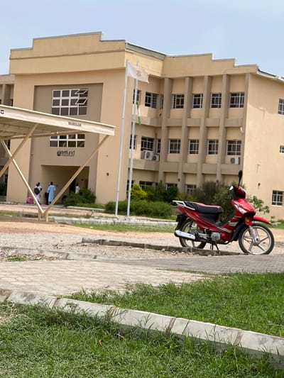 The media file depicts an outdoor scene with a modern, light-yellow, multi-story building in the background. In the foreground, a red and black motorcycle is parked on a paved walkway. To the right of the walkway, a patch of green grass is visible, bordered by a concrete curb. The building features numerous windows, some of which are open. A white banner with a logo is attached to the building near the top. There is a covered walkway in front of the building with metal supports. Some people are visible near the entrance of the building, some appearing to be walking or standing. One person is wearing a light blue robe. Text visible on a sign reads "BURSAR" and a logo with text appears to be for "Tetfund". The scene suggests a daytime setting under clear skies, likely on a university campus or similar institution given the building's architecture and the "Tetfund" branding. The overall atmosphere seems calm and ordinary.