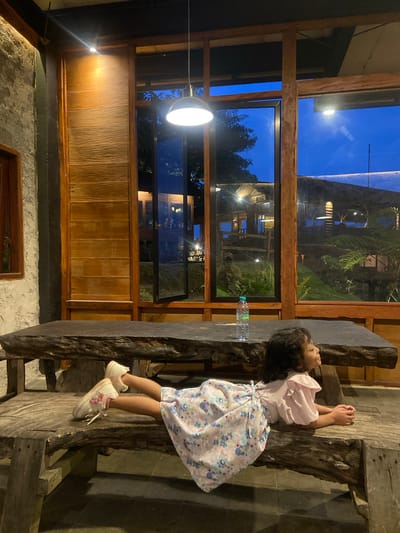 A child in a pink and blue dress is leaning against a wooden table in what appears to be an indoor or partially enclosed space. The table is made of dark wood, and a plastic water bottle is visible on its surface. The background features a large window with a wooden frame, offering a view of a structure and lush greenery in the distance. The lighting suggests it's either nighttime or a dimly lit indoor setting, with an overhead light illuminating the interior. The location is Tamiajeng, Indonesia.