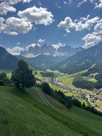 This is a vertical, eye-level, long-shot photo of a scenic mountain village in the Dolomites, Italy. The image is split into three distinct planes. The leftmost third of the image is dominated by a bright blue sky filled with fluffy white cumulus clouds. The middle third showcases a dramatic mountain range with jagged peaks, some snow-capped, receding into the distance. The rightmost third captures a lush green valley with a village nestled within it.
The village, identified as St. Magdalena (Santa Maddalena), is characterized by its cluster of traditional houses with dark roofs, interspersed with green fields and dark green forests. A winding road or path can be seen cutting through the landscape, leading towards the village. In the foreground, on the right, a sloping grassy hill is dotted with small white flowers. A large, leafy tree stands prominently on this hill, with a bench partially visible beneath it.
The scene suggests a beautiful, clear day, likely in the late morning or early afternoon given the bright sunlight casting shadows. The weather appears to be fair and pleasant, ideal for outdoor exploration. The overall mood is serene and picturesque, highlighting the natural beauty and tranquility of the alpine region. No text is visible in the image.