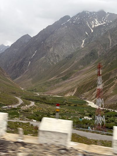 The image depicts a mountain valley in Chikka, India, under an overcast sky. A winding road is visible in the mid-ground, with several vehicles. A prominent red and white lattice tower, likely a telecommunications mast, stands on the right side of the frame. The slopes of the mountains are covered in sparse vegetation and some patches of snow, particularly on the higher peaks. A river flows through the valley floor, bordered by green fields. In the foreground, blurred concrete barriers suggest the image was taken from a moving vehicle on a road. There are no people clearly visible in the scene. The overall environment is rugged and mountainous.