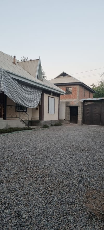 The image shows a residential property on a clear, overcast day. The foreground is dominated by a gravel driveway. To the left, a light-colored house with a dark trim features a corrugated metal roof. A checkered tarp is draped over the front porch area, partially obscuring a window and a wrought-iron railing leading up to the entrance. A metal bench is visible under the porch. Behind this house, a second, larger house made of red brick is visible with a similar corrugated metal roof. This house has multiple windows. In the background, a dark metal gate with a decorative pattern is partially visible. The environment appears to be a suburban or rural setting in the city of Jomi, Tajikistan, suggested by the architectural style and the presence of trees in the background. There are no people or animals visible in the image.