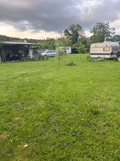 The image depicts an outdoor campsite in what appears to be Dabovets, Bulgaria, characterized by lush green grass. The sky is overcast, suggesting imminent rain. Several caravans and vehicles are present on the site. Under a covered structure, multiple individuals are seated at tables, possibly engaged in dining or socializing. A yellow object is visible amidst the grass, potentially a marker or safety device. The terrain slopes slightly, and various trees dot the landscape.
