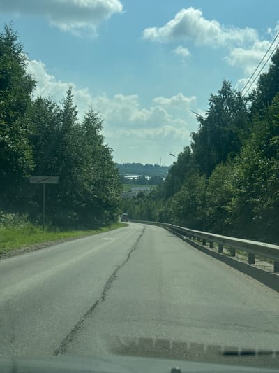 This media file captures a view from a vehicle traveling on a paved road. The road curves to the right, bordered by a metal guardrail on the left and a grassy shoulder. Dense green trees line the left side of the road, creating a canopy that extends into the background. In the distance, beyond the trees, a hazy landscape is visible with hints of industrial structures and a body of water. The sky is a clear blue with scattered white cumulus clouds, suggesting a sunny day. A street lamp is visible among the trees on the left side of the road. A road sign with Cyrillic text, "ГОРОДОК", is partially obscured by foliage on the left. The overall impression is one of a scenic drive through a rural or semi-rural area. The time of day appears to be daytime, given the bright sunlight and clear sky. The weather is fair and pleasant. The activity is the act of driving, as implied by the perspective from inside a vehicle.