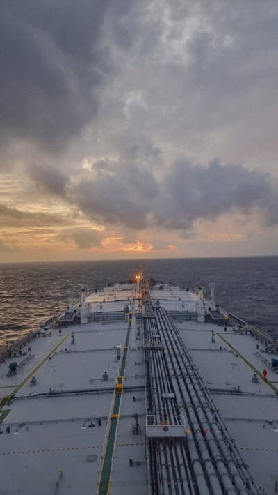 The image shows a large cargo ship sailing on a vast, calm ocean under a cloudy sky at dusk. The sun is low on the horizon, casting a warm glow through breaks in the clouds. The ship's deck is visible in the foreground, with a prominent row of pipes running along its length. The deck is grey and appears to be made of metal. Several yellow and green markings are visible on the deck, likely indicating safety pathways or equipment locations. In the far distance on the right side of the deck, a solitary person in an orange jumpsuit can be seen. The sea is a deep blue-grey, with gentle waves. The sky is a mix of dark, heavy clouds and patches of lighter, more diffused light where the sun is setting. There is no indication of any city or landmass in the image; it is solely a seascape with a ship. The location is not in a city of Unknown, Sri Lanka, as no urban elements are present.