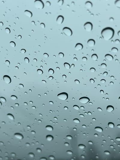 A close-up shot depicts numerous raindrops clinging to a smooth, translucent surface, possibly glass. The drops vary in size, from small to medium, and are uniformly distributed across the frame. Each droplet features a crescent-shaped highlight, indicating a light source positioned above. The background appears blurred and indistinct, contributing to the focus on the water droplets. The color palette is dominated by shades of gray and white, suggesting overcast or indoor lighting conditions.