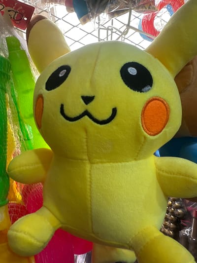 The image shows a close-up of a yellow plush toy, possibly a character like Pikachu, lying on a collection of brightly colored items. The toy's face is the focal point, with a large black eye on the left side, a stitched black mouth, and an orange circle cheek on the right. Its body is soft and fuzzy, with visible stitching.
The plush toy is positioned above a mesh-like material, under which are several objects. Some appear to be plastic toys or containers, in shades of green, pink, and possibly red. The background is blurred but suggests a retail or market environment, with other items and possibly a wire rack visible.
There is a red tag hanging from the lower left corner of the plush toy. Visible text on the tag includes "dear" and possibly "bol bol" or similar short words, along with a small drawing of a person.
The lighting appears to be natural daylight, casting soft shadows and highlighting the textures of the plush toy. There are no people clearly visible in the image. The setting suggests a shop or a stall, likely in Tokmok City, Kyrgyzstan, given the location context. The overall impression is of a playful and colorful display of merchandise.