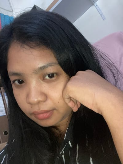 A young woman with long, black hair rests her face on her hand. She has brown eyes, and her skin tone is medium. She is wearing a black and white striped shirt. The location is Negara, Indonesia, indicated by the presence of a shipping box with associated labels commonly found in Indonesia, but not explicitly visible. Other objects in the scene include a pink surface in the background and stacked items on a surface. The image is taken from a close-up, slightly tilted perspective.