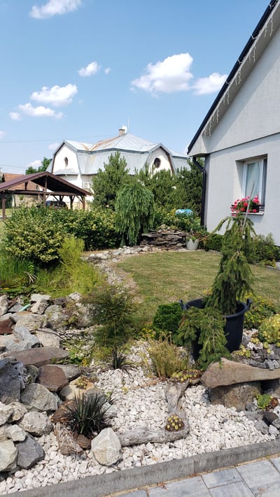 This media file depicts an outdoor scene with a building, garden, and sky. The building appears to be a church or similar structure with a white exterior and a silver, multi-peaked roof. It has round windows and is partially obscured by greenery. Attached to the building, or a separate structure nearby, is a covered patio area with a brown roof supported by wooden beams.
The garden is extensive and features a variety of plants, including evergreens, bushes, and ground cover. There is a grassy area, and the ground is covered in white gravel and large rocks of various sizes and colors, forming a decorative rock garden. Some plants, possibly succulents, are growing in a piece of driftwood in the rock garden. A black plastic planter is also visible. A window on an adjacent white building has a planter box with pink flowers.
The sky is a clear blue with scattered white clouds, suggesting daytime and fair weather. The scene is likely a residential or public area in Vyšné Nemecké, Slovakia, given the location context. There are no people visible, and the overall impression is one of a well-maintained garden and building. The image is oriented vertically, and the perspective is from a low angle, looking upwards towards the sky and the structures. There is no visible text in the image.