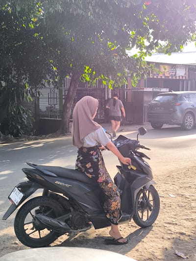 The photo shows a woman on a black Honda motorcycle, stationary on a street in Indonesia. She is wearing a brown hijab, a light-colored shirt, and floral print pants, and is holding a key in the ignition. The motorcycle has a license plate that reads "DR 6435 UV." In the background, a person walks toward a building with the signage "GGSP IDENTITAS GUE SIGNATURE! TOKDATOR." A dark gray Mazda CX-5 with license plate "B 2263 UZI" is parked nearby. The environment includes trees, a fence, and a building identified by the signs, suggesting this is a commercial area or a small business location in Selongbelanak, Indonesia. The image is taken during the daytime with bright sunlight creating shadows.