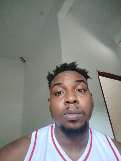 The image is a close-up, forward-facing selfie of a Black man wearing a white tank top with red trim. He has short, dark, curly hair and a goatee. The background is a wall and a ceiling with a subtle shadow cast by a light source. There is a small, dark object on the ceiling. The man's facial expression is neutral. The image does not provide any information about the location in the city of Lahat, Indonesia.