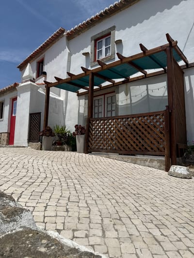 The image shows a white building with a red-tiled roof and red-trimmed windows, likely a house or a small establishment, situated on a cobbled street in the town of Colares, Portugal. A wooden pergola with a green mesh cover extends from the building, providing shade over a patio area. The patio is enclosed by a wooden lattice fence. Two large white planters with succulent plants are placed near the entrance. The street is paved with light-colored cobblestones that slope upwards. The sky is clear blue with some wispy clouds. The scene is bathed in bright sunlight, creating strong shadows. There are no people visible in the image.