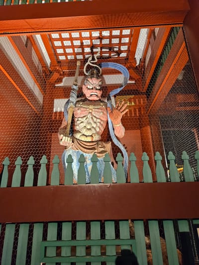 The image shows a statue of a multi-armed deity behind a wire mesh. The statue is predominantly reddish-pink and gold in color. It has a mask-like face and is surrounded by blue and white ornaments. The statue is located inside a structure with an orange lattice on one side and a green structure with rounded edges on the other. The location is likely in Taito, Japan, given the statue's style and cultural context.