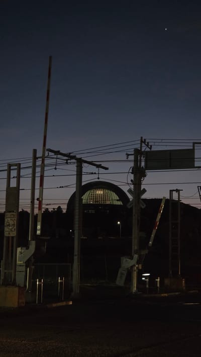 The image shows a railroad crossing at night, with crossing arms and signage. In the background is a large, arched structure with a grid-like pattern, illuminated from within. Above and to the left, a single, bright celestial object (likely a star or planet) is visible in the night sky. The environment appears to be urban, with utility poles and power lines visible. The location is Guadalajara, Mexico.