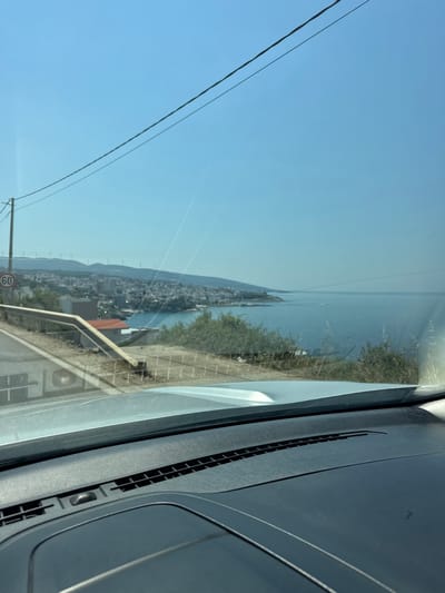 The image, taken from inside a vehicle, shows a scenic view of the coastline and a town. In the foreground, the dashboard of the car is visible, with a gray textured surface and air vents. Through the windshield, a road curves to the left, bordered by a metal guardrail. A speed limit sign indicating "60" is mounted on a pole on the side of the road.

Beyond the road, a hillside slopes down towards the sea. On the hillside, there are several buildings, some with red roofs, and dense green vegetation. In the middle ground, a coastal town extends along the bay, with numerous buildings and structures. A body of calm, blue water forms the bay, with a hazy horizon in the distance. On the distant hills, several wind turbines are visible, suggesting a potentially windy location. The sky is clear and blue.

The image captures a daytime view of a coastal town, likely Dobra Voda, Montenegro, characterized by its proximity to the sea, hilly terrain, and the presence of modern infrastructure like wind turbines. The scene appears tranquil, with no people or vehicles immediately discernible in the outdoor portion of the image.