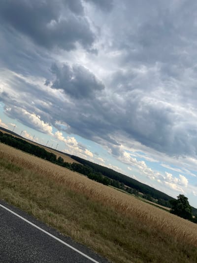 This image shows a landscape view of a field of wheat with a dark grey asphalt road in the foreground. The field extends into the distance and is bordered by a dense line of dark green trees. Beyond the trees, several wind turbines are visible against a sky filled with dramatic, dark grey clouds interspersed with patches of blue sky and white clouds. The overall scene suggests a rural setting, likely in the vicinity of Holzburg, Germany, characterized by agricultural land and renewable energy infrastructure. There are no people or animals visible in the image. The lighting suggests it is late afternoon or early evening, with the heavy cloud cover creating a moody atmosphere.