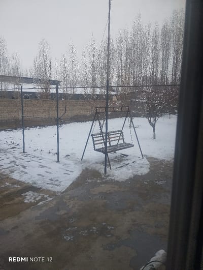 A wintery scene features a swing set in a yard with patchy snow cover. Bare trees stand in the background, with a wall and building partially visible behind them. The foreground is a concrete or stone surface with melting snow. The photo is taken from indoors, with a doorframe visible on the right. The overall atmosphere is cold and overcast.