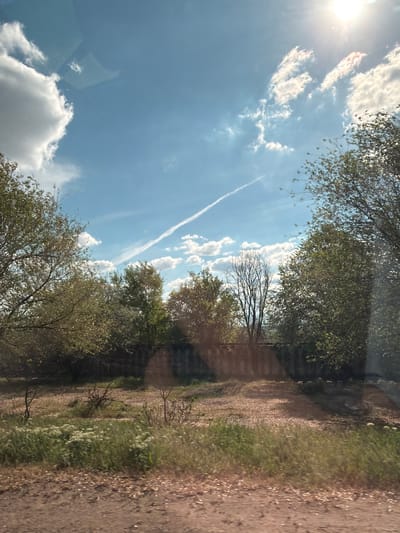 The image shows a rural scene in Taraclia, Moldova. A dirt road runs along the right side of the frame. The left side of the image is dominated by a bright blue sky with scattered cumulus clouds and a contrail. Trees line both sides of the road. The image appears to have been taken from inside a vehicle, with a faint reflection visible in the lower left corner. There are no people visible.
