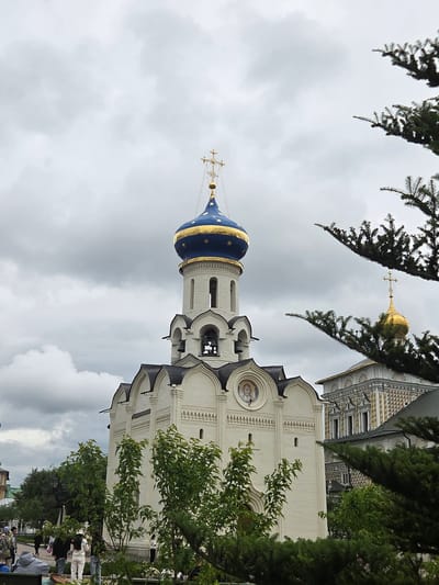 The image shows a white building with a blue and gold dome, topped with a cross, in the city of Sergiev Posad, Russia. The building appears to be a church or cathedral. Several people are visible in the foreground and middle ground, some walking and some standing. They are dressed in casual clothing. Evergreen trees are present on the right side of the image, partially obscuring the view of the buildings. The sky is overcast with gray clouds. In the background, another building with a golden dome and ornate architectural details is visible.