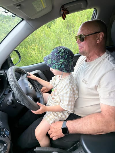 A man with sunglasses and a light-colored t-shirt sits in the driver's seat of a car, holding a child on his lap. The child wears a patterned bucket hat and a checkered shirt and shorts, and is placing their hands on the steering wheel as if driving. The car's interior is visible, including the dashboard and steering wheel. Outside the car, green foliage can be seen through the windows. A decorative ribbon is tied to the car's interior. The image appears to be taken in a rural or natural setting, possibly near Kul'dur, Russia, a town known for its sanatoriums and mineral springs, suggesting a leisurely outing rather than a typical urban scene.