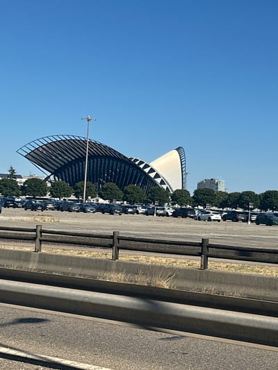 The image captures a wide view of a modern architectural structure, likely a stadium or arena, with a distinctive curved, wing-like roof made of a light-colored material and a dark, lattice-like support system. The building is set against a clear, bright blue sky, indicating a sunny day. In the foreground, a paved area, possibly a parking lot, is visible, with several cars parked in neat rows. Trees line the edge of the parking lot, separating it from the building. A tall light pole stands near the structure.
To the right of the scene, a metal guardrail runs along what appears to be a road or highway, with asphalt paving. The guardrail is a prominent feature in the right half of the image, composed of vertical posts and horizontal beams. The overall impression is of a public space, possibly on the outskirts of a city, designed for large gatherings or events. The presence of a parking lot suggests that the location is meant to accommodate visitors. The clear sky and bright sunlight suggest it is daytime, likely midday or early afternoon. No people are visible in the frame. The location context is Colombier-Saugnieu, France.