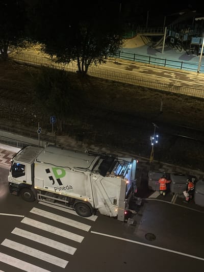 The image depicts a nighttime scene in Pineda de Mar, Spain, captured from an elevated perspective. A white and light green rear-loader garbage truck, emblazoned with "Pineda a punt," is stationary on a wet asphalt road. The front half of the truck rests on a white-striped pedestrian crosswalk, while its rear is positioned adjacent to multiple waste containers on the curb.

Two individuals, identifiable as waste management personnel, are present near the rear of the truck. Both wear orange high-visibility jackets. One individual stands upright near a large, rectangular, grey waste bin, appearing to operate or prepare it for collection. The second individual is bent over a smaller, dome-lidded white and blue bin, possibly handling its contents. The rear of the garbage truck emits bright white light, illuminating the immediate collection area.

The environment includes a tiled sidewalk to the left of the road, leading to a sloped, vegetated embankment. Below this embankment, train tracks are visible. Traffic signs, including a pedestrian crossing sign and a blue circular sign, are present near the tracks. Across from the waste collection operation, separated by a pedestrian path and a fence, lies a brightly lit children's playground featuring slides, climbing structures, and a ship-like play area on a blue surface. The entire scene is illuminated by streetlights and the truck's operational lights, indicating ongoing activity.