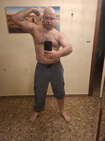 A shirtless, bald man stands in an interior space, likely a bathroom, as suggested by the presence of a mirror. He's wearing grey pants and is holding a black smartphone in his right hand, obscuring part of his torso. He is flexing his left bicep. The man appears to be positioned in front of a wall with a painting of a cityscape hanging on the left side, next to the light switch and house keys. The floor is tiled with a speckled brown pattern. The location could be Xirivella, Spain, as implied in the instructions, though specific details confirming this are not visually present.