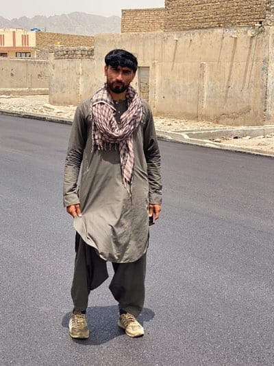A man stands on a freshly paved road in what appears to be قندهار, Afghanistan. He wears a traditional gray shalwar kameez and a checkered scarf around his neck, with dirty athletic shoes. The background reveals unfinished brick buildings and a barren, mountainous landscape.
