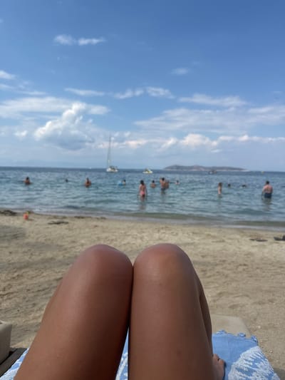In Thassos, Greece, on a sunny day, a close-up shot focuses on the thighs of a person lying on a beach chair with a blue and white striped towel. Beyond the person's legs, the sandy beach extends to the edge of the calm, blue sea. Several people are visible in the water, some swimming and others standing. A sailboat with a white sail is anchored in the distance, and a hazy island can be seen on the horizon under a sky filled with scattered white clouds. The scene conveys a relaxed, summery atmosphere at a seaside resort.
