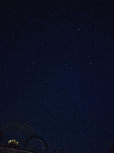 The image captures a dark, starry night sky with a few visible stars. In the lower left quadrant, there is a dark, ornate metal structure that appears to be part of a gate or fence. This structure has decorative curves and a small, golden, knob-like element is visible. The background is a deep navy blue, indicative of nighttime. The scene is likely set in Schwangau, Germany, given the context provided, possibly near one of its famous castles like Neuschwanstein or Hohenschwangau, suggesting an outdoor setting at night.