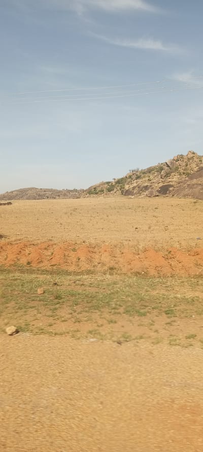 The image displays a broad outdoor landscape under a pale blue sky with faint, wispy clouds, suggesting clear daytime conditions. The foreground features dry, sandy soil interspersed with patches of sparse green grass. A prominent horizontal band of reddish-brown earth with small mounds, possibly termite hills or eroded soil, crosses the mid-foreground. Beyond this, a wide expanse of dry, yellowish-brown land extends towards the horizon, indicating an arid or dry environment.

In the background, a series of low, rocky hills and outcrops rise, covered with scattered grey and brown boulders and sparse green vegetation, including shrubs or small trees. Three thin, white power lines are visible stretching horizontally across the upper portion of the sky, hinting at nearby infrastructure. No human activity, people, or discernible objects other than the natural landscape and power lines are visible. The overall scene conveys a sense of rugged natural beauty and openness typical of a dry, sunny region, consistent with the specified location of Barakin Kuru III, Nigeria. There is no visible text.