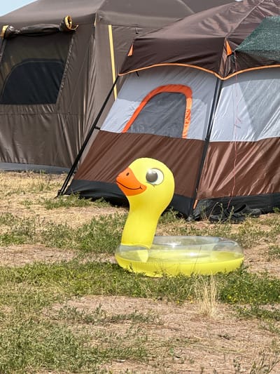 The image shows a yellow inflatable duck toy with a clear ring float in front of a large tent. The tent is dark brown with orange trim and has a window on the left side. The ground is covered in dry grass and some scattered green vegetation. There are a pair of black shoes visible near the base of the tent. The scene appears to be outdoors, possibly in a campground or a rural area. The lighting suggests it is daytime. The specific location in the city of Kolodezny, Russia cannot be determined from the visual information alone.