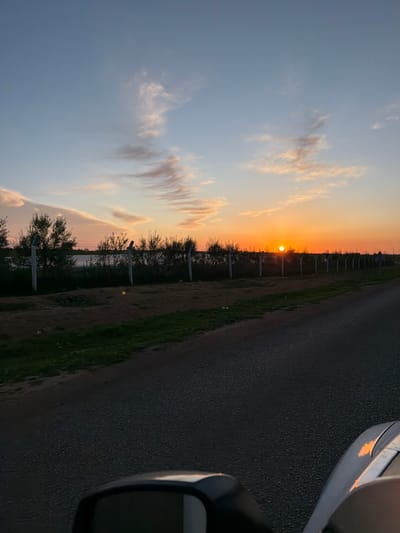 This image depicts an outdoor, roadside scene in Sabaa Aiyoun, Morocco, at sunset. The bright orange sun is visible as a distinct orb positioned just above the horizon in the upper-right quadrant of the image. The sky transitions from a light blue at the zenith to vibrant orange and pink hues near the horizon, with elongated, wispy clouds reflecting these warm tones.

In the foreground and lower-midground, a dark asphalt road runs diagonally from the lower-left towards the right side of the frame. To the immediate left of the road, a narrow strip of green grass is visible, followed by a wider expanse of reddish-brown dirt. A fence parallels the road on the left, composed of numerous light-colored posts with darker caps, and what appears to be a chain-link or barbed-wire section running between them. Behind this fence, a dense line of dark green shrubs and small trees stretches across the midground. Beyond the vegetation, a lighter, indistinct band extends horizontally, possibly indicating a distant structure or body of water.

In the lower-right corner, the front fender and a portion of the side mirror of a light-colored vehicle are visible, suggesting the photograph was taken from inside or beside a moving vehicle. The vehicle's surface reflects the warm orange light of the setting sun. No individuals or specific actions are discernible within the frame.