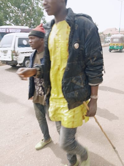 An outdoor street scene in Kwekwe, Zimbabwe, depicts two young males walking on a paved road under bright daylight. In the foreground, a male with dark skin wears a bright yellow textured tunic beneath a dark, possibly black, puffy jacket bearing a circular pin. He wears dark trousers and light green slip-on shoes. His right hand grips a slender wooden stick pointed downwards, and his left wrist is adorned with multiple light-colored beaded bracelets. His face is partially obscured. Slightly behind and to his left, a second male with dark skin wears a red and black knitted beanie hat, a dark open jacket over a light brown t-shirt with a graphic design, dark trousers, and matching light green slip-on shoes. He looks towards the left side of the frame, with his right arm bent at the elbow. Both individuals show motion blur, indicating movement. In the background, a white minibus, partially obscured, displays "RAISE & WORSHIP" text on its side. Further down the road on the right, a green and yellow auto-rickshaw is visible, moving away. Trees line the distant background, and faint building structures are discernible under a bright sky.