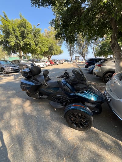 A three-wheeled motorcycle, a dark teal Can-Am Spyder F3-S, is parked on a paved surface in what appears to be a parking area near a beach. The motorcycle is positioned in the foreground, angled slightly towards the right. Behind it, a row of parked cars can be seen, including a silver Lexus SUV, a dark blue SUV, and various other passenger vehicles. Trees with green foliage line the background, with their branches casting shadows on the ground. In the far distance, a glimpse of the sea and a few people on the beach are visible under a clear blue sky. The overall scene suggests a sunny day in a coastal town. The license plate of the silver Lexus SUV is visible and reads "TMB 765".