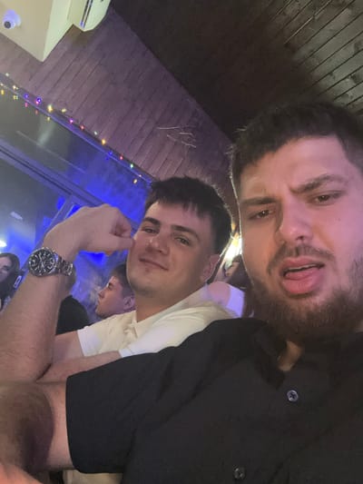 The image shows a selfie of two young men, possibly in a bar or club setting. The man in the foreground is wearing a white collared shirt and a watch on his left wrist. Behind him, another man is partially visible, appearing to wear a black shirt. The man at the top of the image is sticking his tongue out. The background shows dimly lit surroundings with colored lights and what appears to be a wooden ceiling. A security camera is visible on the top left of the image. It is not possible to determine if the location is Panagyurski kolonii, Bulgaria, from the visual information provided.